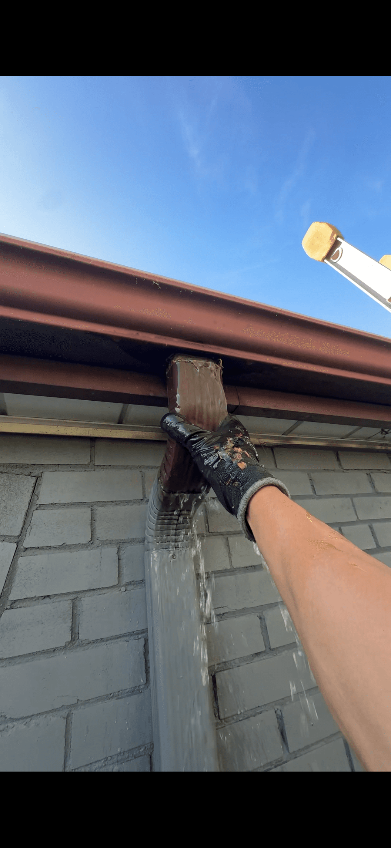 Gloved hand inspecting a leaking brown gutter downspout overflowing with water on a brick wall.