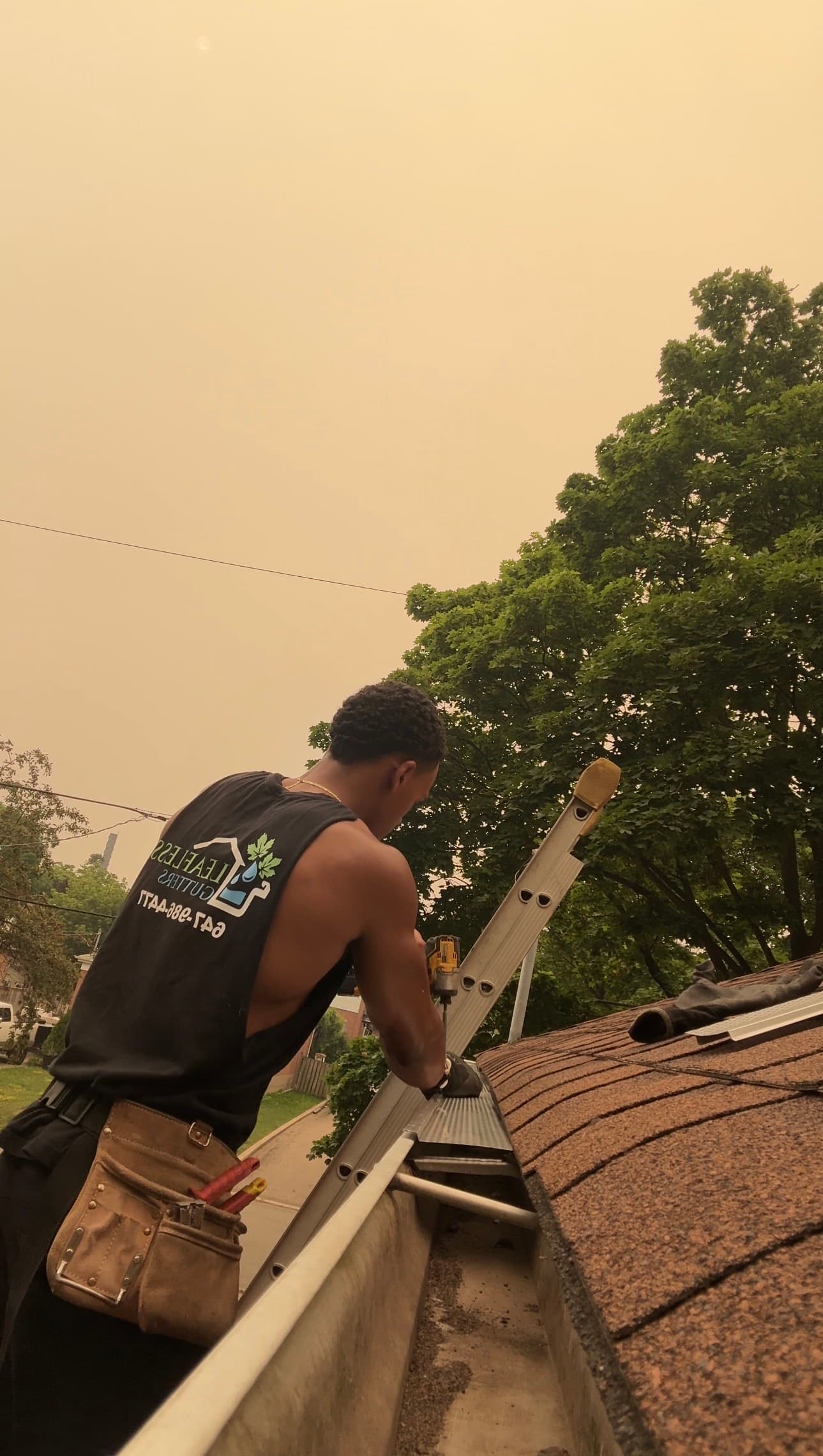 A man installs gutter guards on a roof under a hazy, orange-tinted sky.