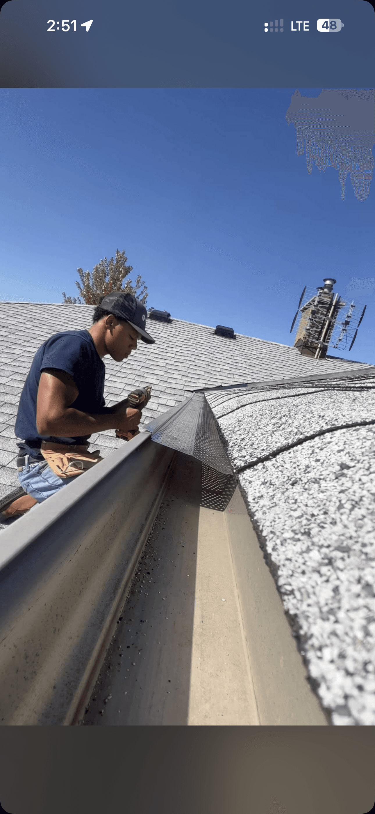 Man installing metal mesh gutter guards on a shingled roof using a power drill.